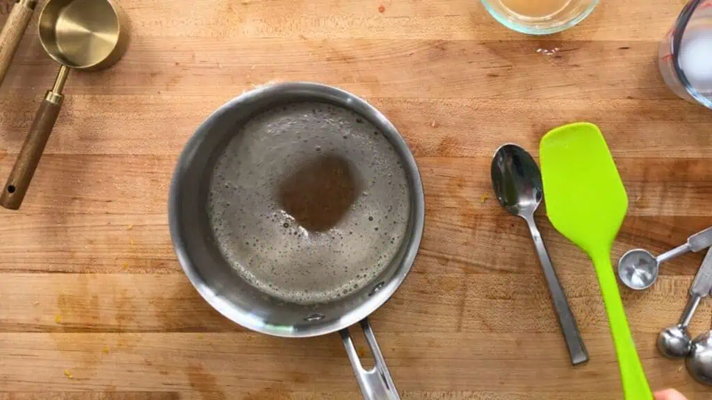A saucepan with bubbling liquid sits on a wooden countertop beside a metal spoon, a green spatula, measuring cups, and a glass bowl.