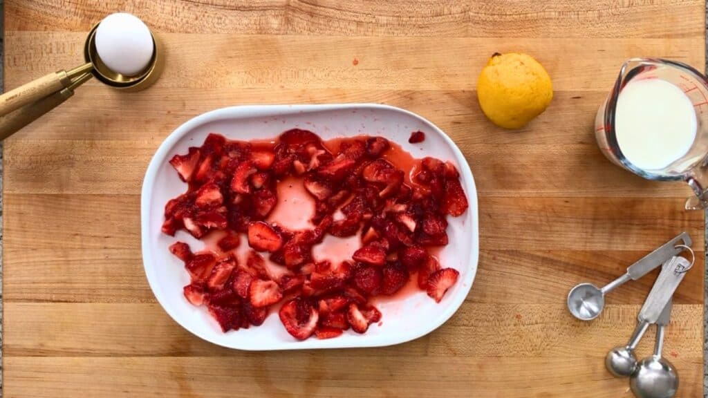 Sliced strawberries in a white dish on a wooden surface, with an egg, a lemon, a measuring cup of milk, and metal measuring spoons nearby.