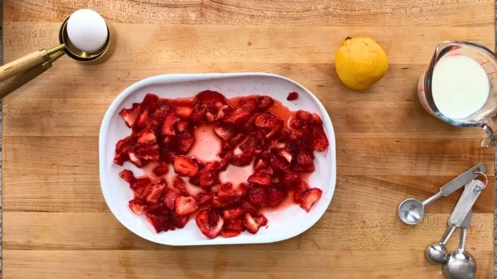 Sliced strawberries in a white dish on a wooden surface, with an egg, a lemon, a measuring cup of milk, and metal measuring spoons nearby.