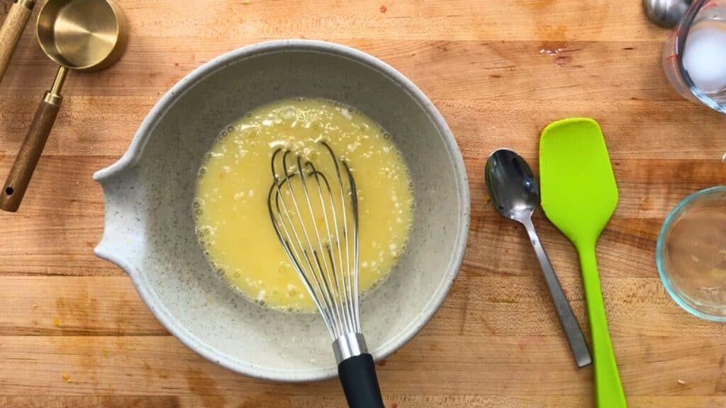 A whisk is mixing beaten eggs in a ceramic bowl on a wooden countertop, with a spoon, a green spatula, measuring cups, and an empty glass nearby.