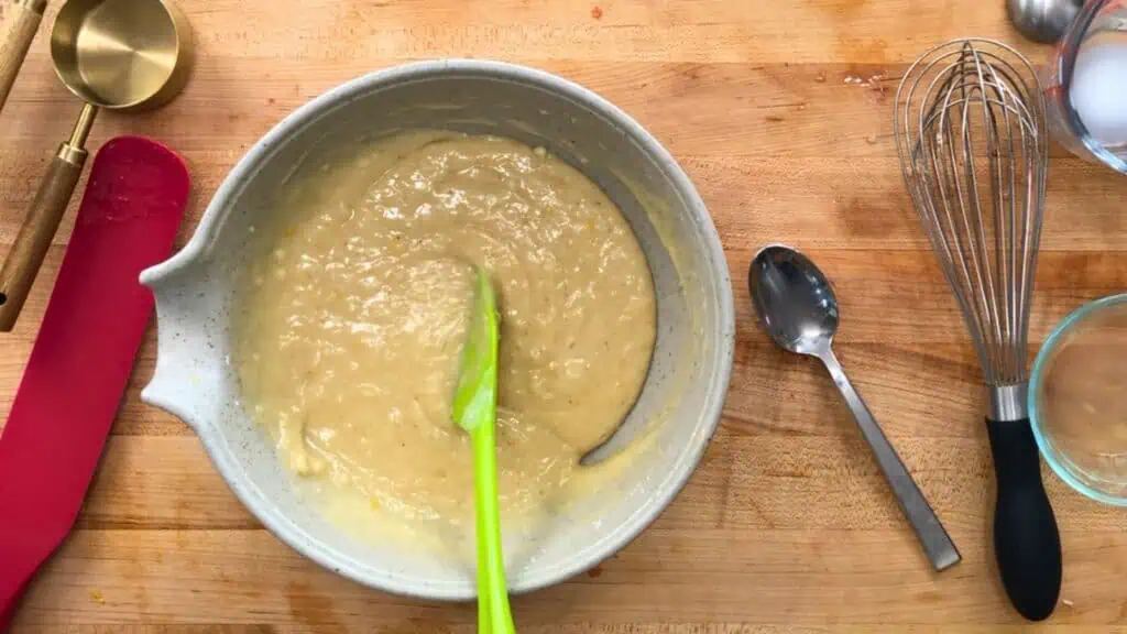 A mixing bowl with batter and a green spatula on a wooden surface, surrounded by a spoon, whisk, measuring cup, and other baking tools.