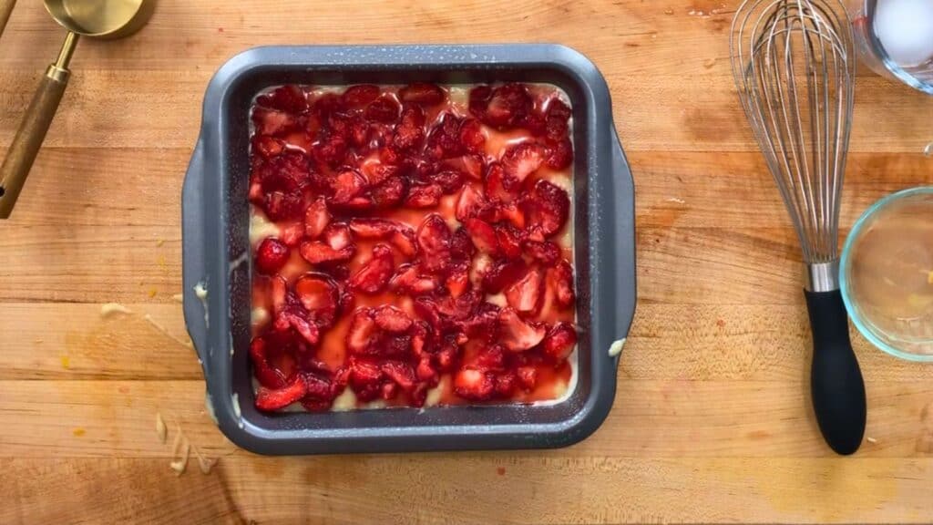 A square baking pan filled with an unbaked dessert topped with sliced strawberries sits on a wooden countertop, surrounded by baking tools and ingredients.