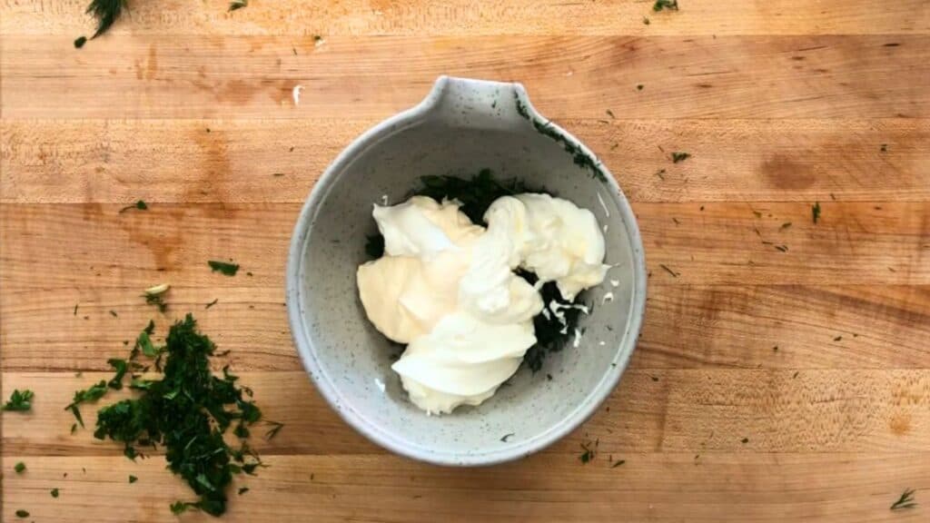 A gray bowl containing chopped herbs, mayonnaise, and sour cream sits on a wooden surface sprinkled with additional herb pieces.