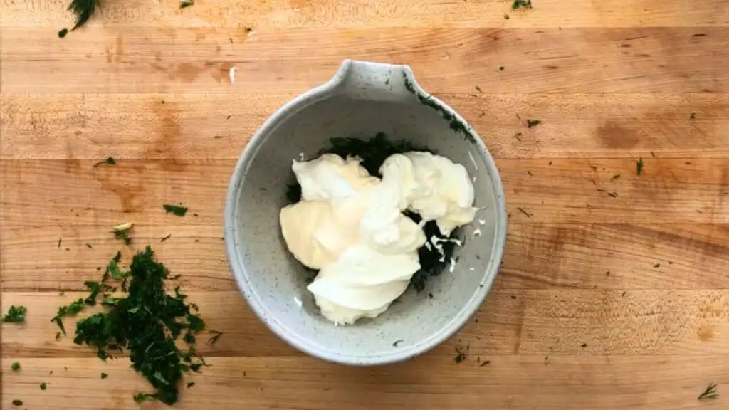 A gray bowl containing chopped herbs, mayonnaise, and sour cream sits on a wooden surface sprinkled with additional herb pieces.