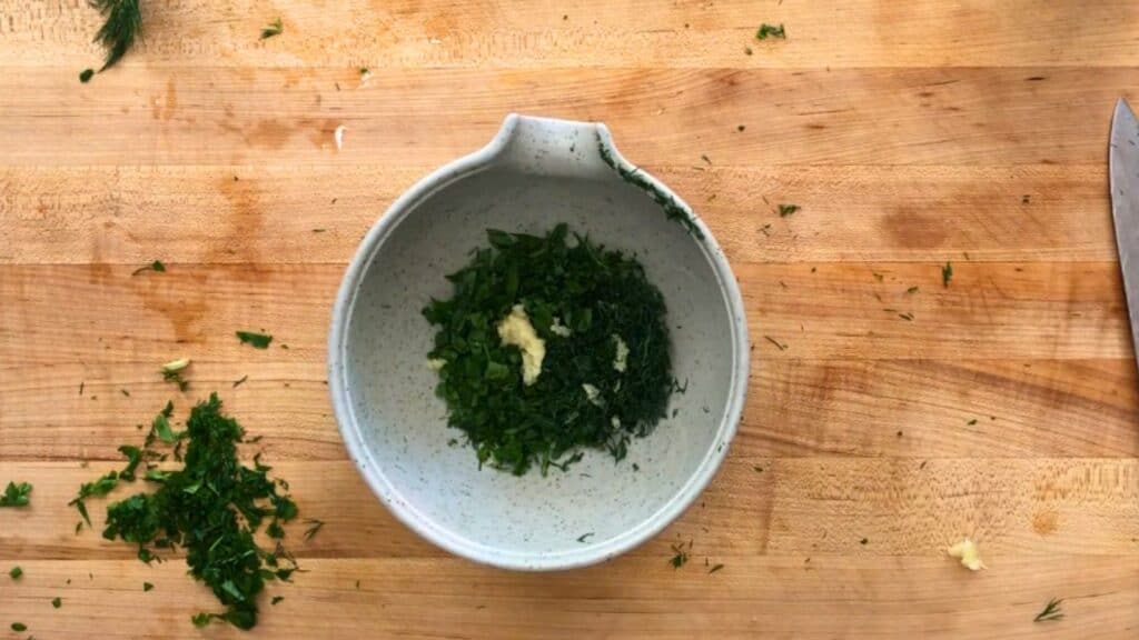 Chopped fresh dill and parsley with minced garlic in a speckled white bowl on a wooden cutting board.