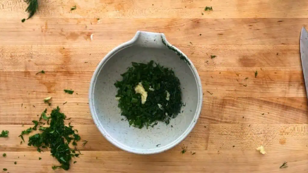 Chopped fresh dill and parsley with minced garlic in a speckled white bowl on a wooden cutting board.