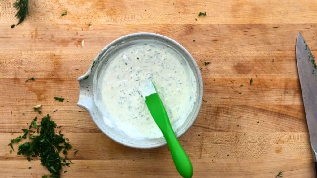 A bowl of creamy herb sauce with a green spatula sits on a wooden cutting board, surrounded by chopped herbs and a large kitchen knife.
