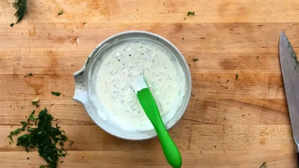 A bowl of creamy herb sauce with a green spatula sits on a wooden cutting board, surrounded by chopped herbs and a large kitchen knife.