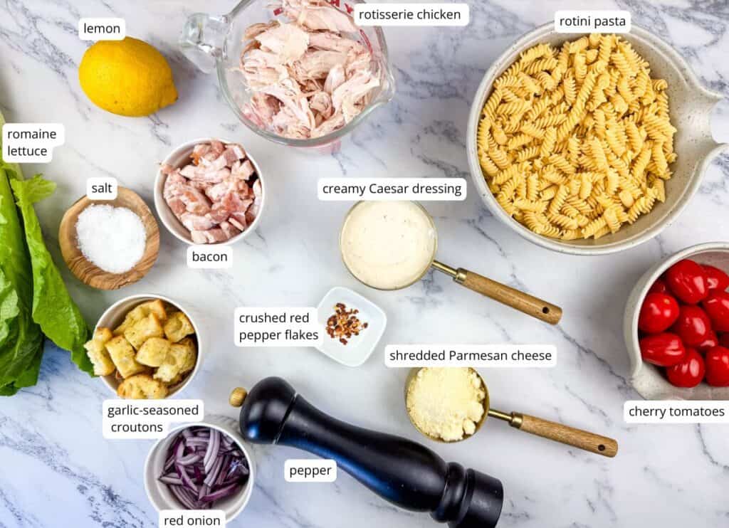 Ingredients for a rotini pasta salad are laid out on a counter, including pasta, rotisserie chicken, Caesar dressing, bacon, cheese, lettuce, tomatoes, croutons, onion, lemon, salt, and pepper.
