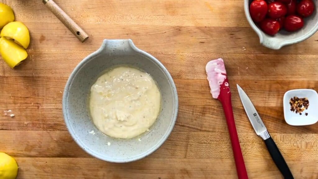 A bowl of batter sits on a wooden countertop, surrounded by a spatula, a knife, lemon wedges, a bowl of tomatoes, and red pepper flakes.