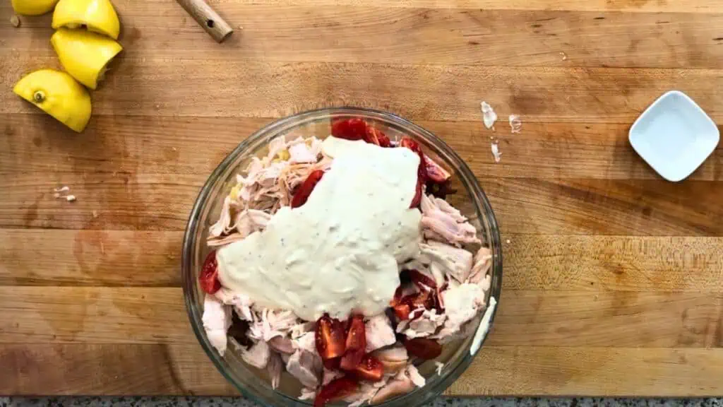 A glass bowl containing shredded chicken, chopped tomatoes, and a dollop of creamy dressing sits on a wooden cutting board with lemon wedges and a small dish nearby.