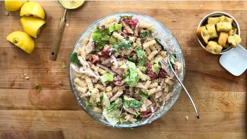 A glass bowl of pasta salad with rotini, lettuce, tomatoes, and dressing sits on a wooden surface with a spoon, lemon wedges, croutons, and a measuring spoon nearby.