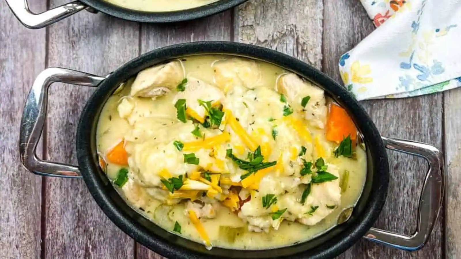 Oval bowl of chicken and dumplings in creamy sauce, topped with shredded cheddar cheese and chopped parsley, on a wooden table next to a floral napkin.