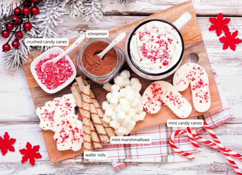 A wooden board with mugs of hot chocolate, mini marshmallows, wafer rolls, labeled jars of crushed candy canes and cinnamon, and mini candy canes, on a white rustic table.