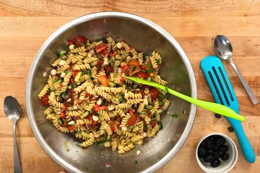 A large metal bowl of pasta salad with rotini, cherry tomatoes, red onion, and herbs sits on a wooden surface. Utensils, a bowl of black olives, and a blue spatula are nearby.