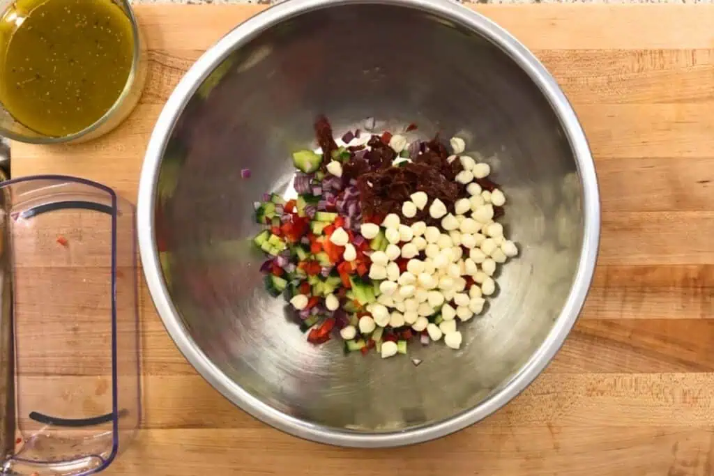 A metal mixing bowl contains diced red onion, cucumber, red and green bell pepper, sun-dried tomatoes, and white cheese chips. A small dish of vinaigrette sits nearby on a wooden surface.