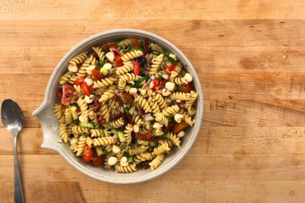A bowl of rotini pasta salad with cherry tomatoes, mozzarella balls, red onion, and herbs on a wooden surface next to a spoon.