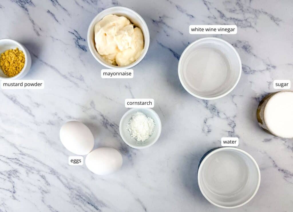 Ingredients on a marble surface: mustard powder, eggs, cornstarch, mayonnaise, white wine vinegar, sugar, and water, each in separate bowls and labeled.