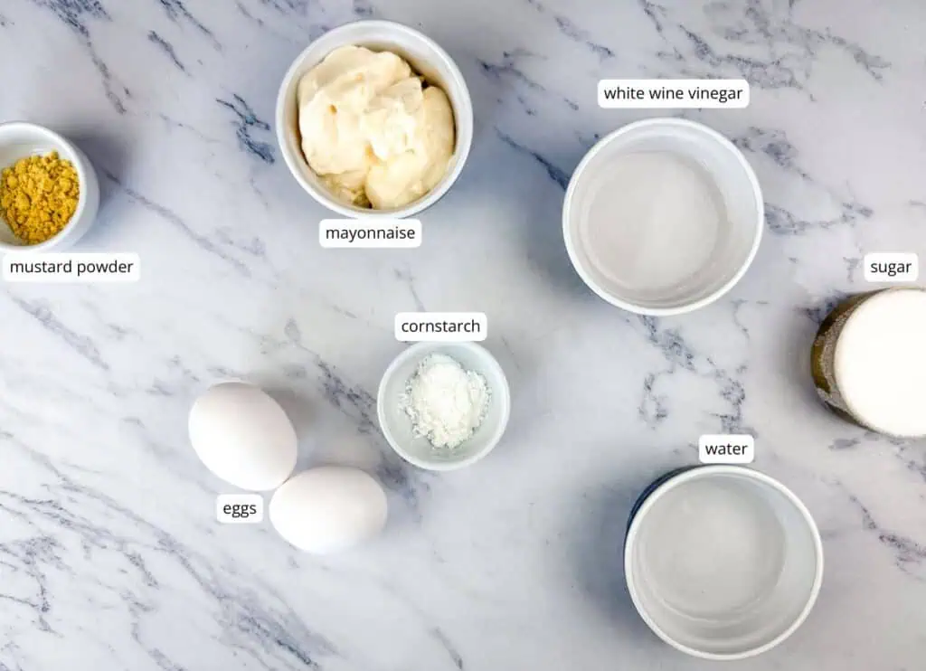 Ingredients on a marble surface: mustard powder, eggs, cornstarch, mayonnaise, white wine vinegar, sugar, and water, each in separate bowls and labeled.