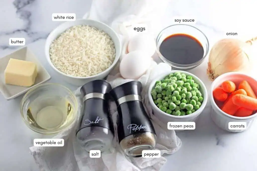 Ingredients for fried rice, including white rice, eggs, soy sauce, onion, butter, vegetable oil, salt, pepper, frozen peas, and carrots, arranged on a white surface with labels.