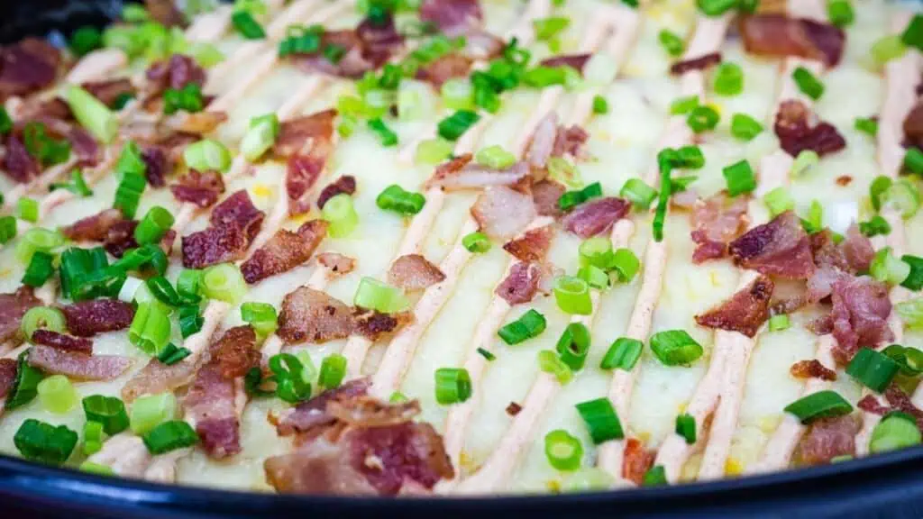 Close-up of a baked dish topped with chopped bacon, sliced green onions, and a drizzle of sauce arranged in parallel lines.