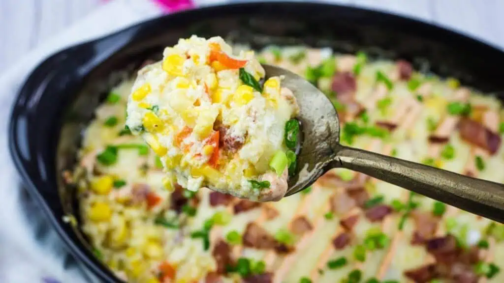 A spoon holds a serving of creamy casserole with corn, chopped vegetables, and green onions above a baking dish filled with the same dish.