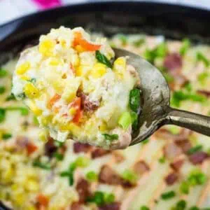 A spoon holds a serving of creamy casserole with corn, chopped vegetables, and green onions above a baking dish filled with the same dish.