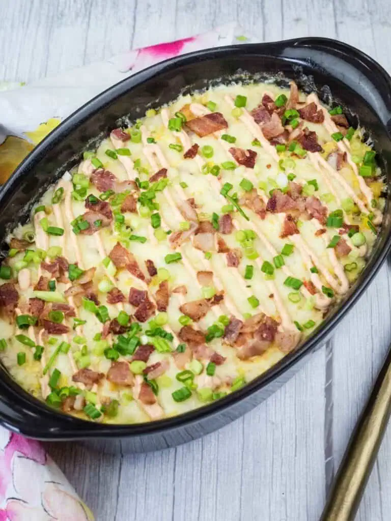 A black oval dish filled with baked mashed potato casserole, topped with chopped bacon, green onions, and lines of melted cheese, sits on a wooden surface.
