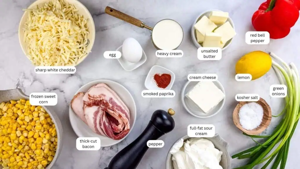 Ingredients for a recipe are arranged on a marble surface, including cheeses, bacon, corn, egg, heavy cream, sour cream, butter, spices, red bell pepper, lemon, green onions, and seasoning.