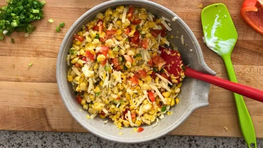 A bowl of corn salad with chopped red bell pepper, green onions, and shredded cheese on a wooden cutting board with a green spatula and chopped vegetables nearby.