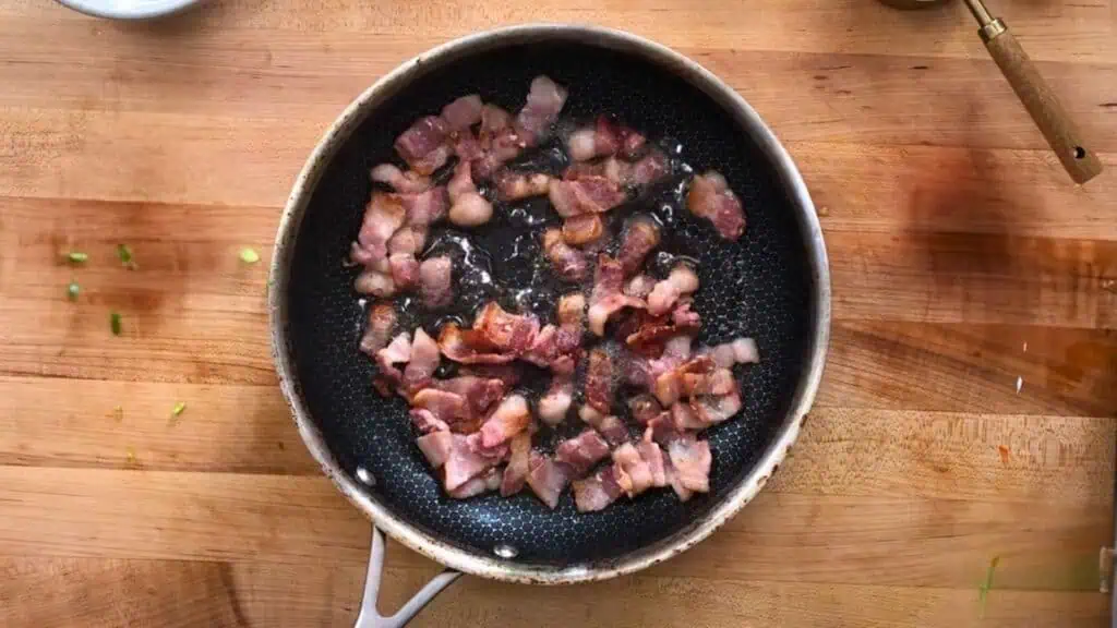 A frying pan with pieces of bacon cooking on a wooden countertop.