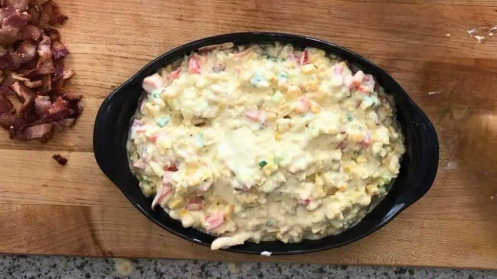 A black oval dish filled with a creamy mixture of corn, chopped vegetables, and cheese sits on a wooden cutting board next to crumbled bacon.