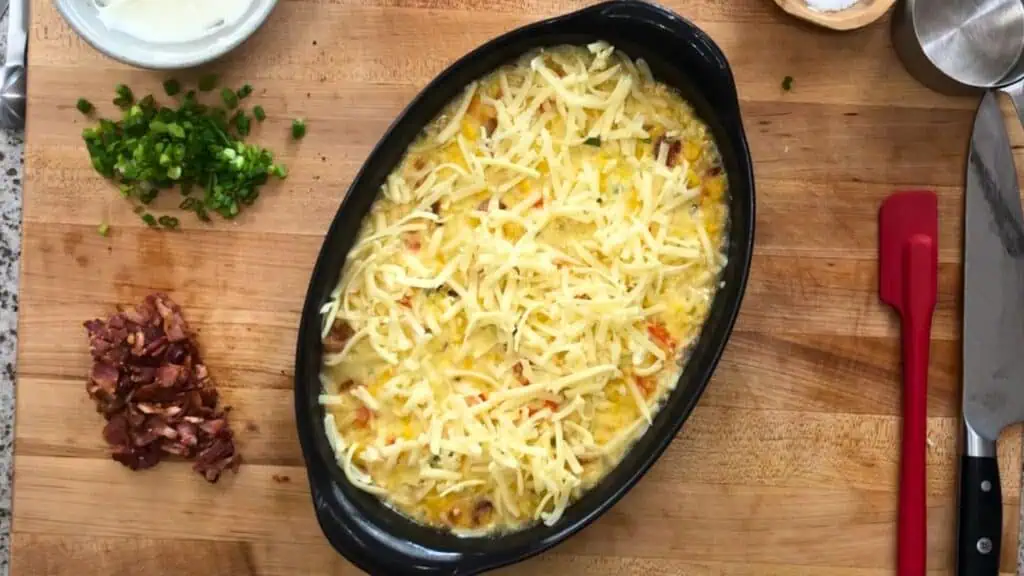 Oval baking dish filled with a cheese-topped casserole sits on a wooden board, surrounded by chopped bacon, green onions, a knife, and small bowls.