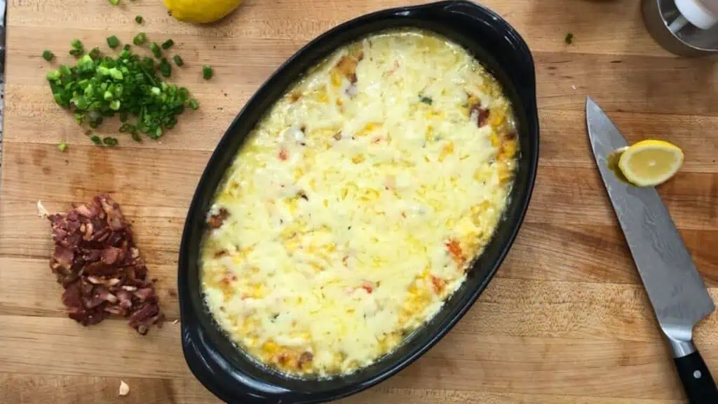 Oval baking dish with cheesy casserole on a wooden surface, surrounded by chopped green onions, bacon, a knife, lemon wedge, and a salt grinder.