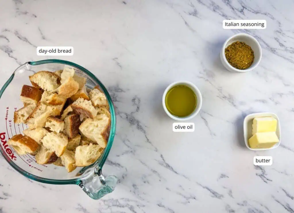 Cubed day-old bread in a measuring cup, with small bowls of Italian seasoning, olive oil, and butter arranged on a marble surface.