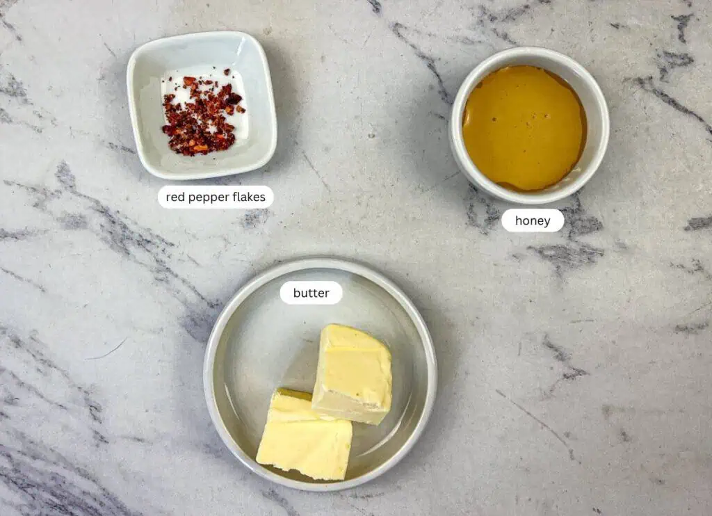 Three bowls on a marble surface, containing red pepper flakes, honey, and two pieces of butter, each labeled accordingly.