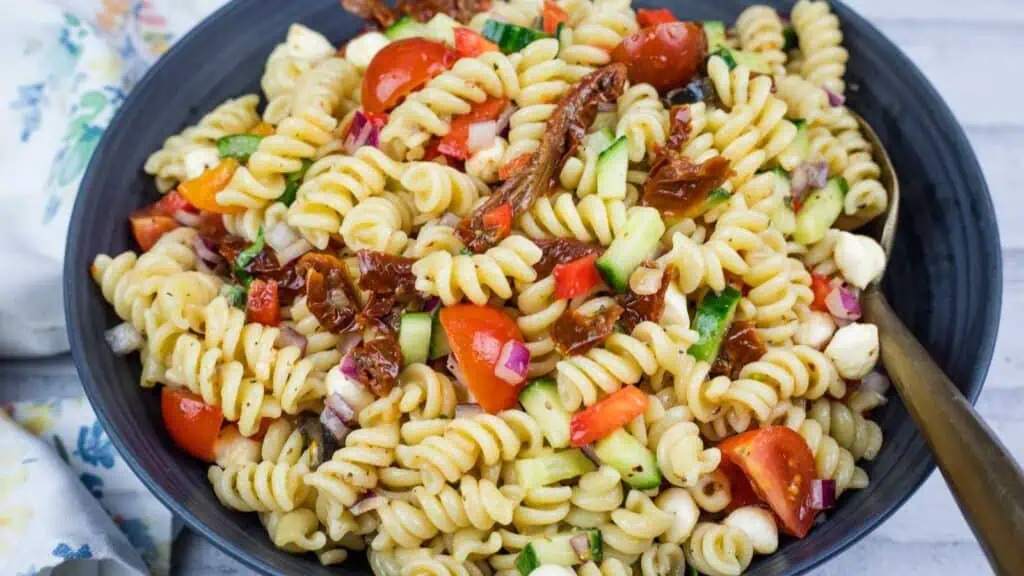 A bowl of Italian pasta salad with cherry tomatoes, cucumber, red onion, mozzarella balls, and sun-dried tomatoes, served with a fork.
