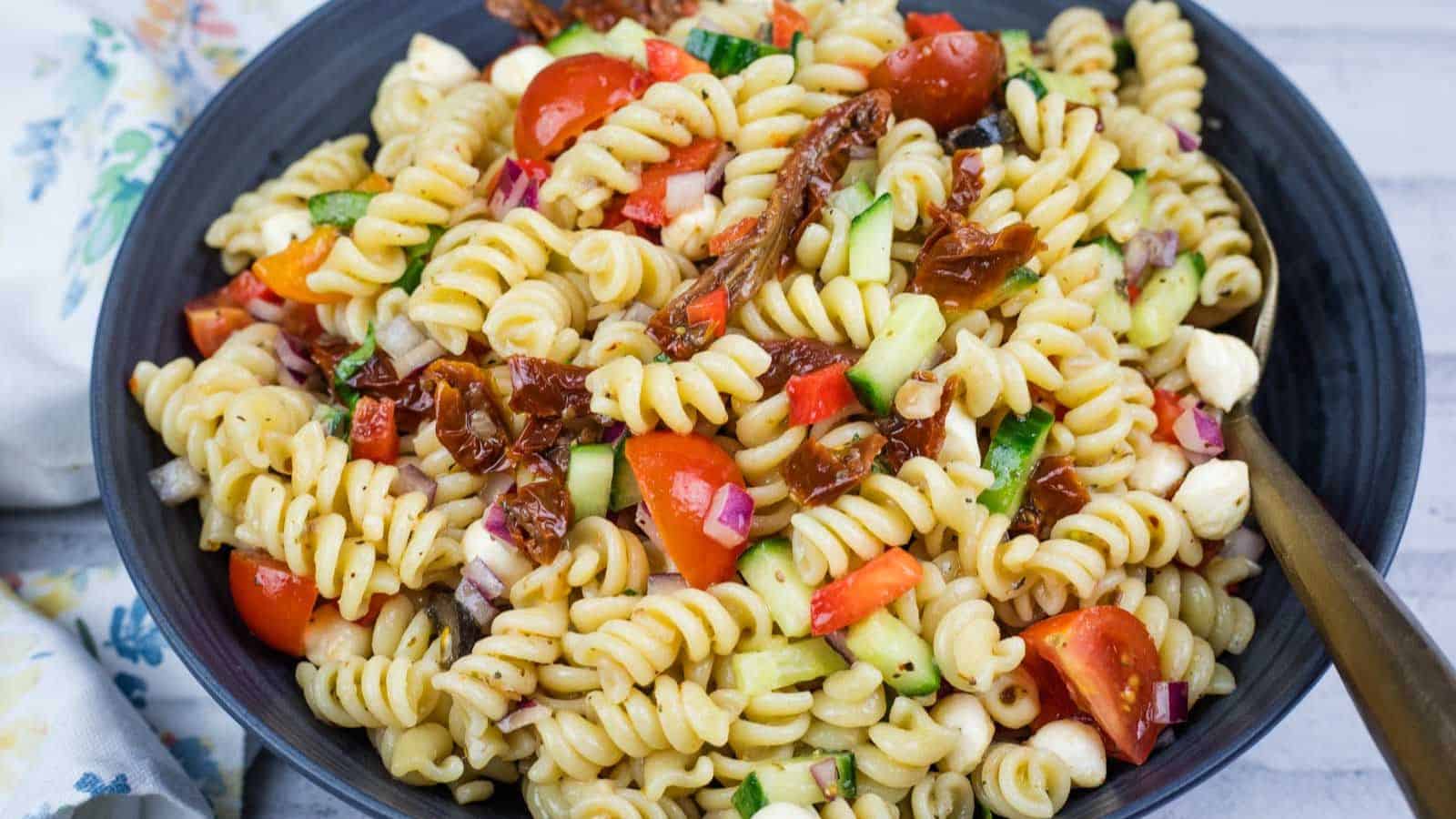 A bowl of Italian pasta salad with cherry tomatoes, cucumber, red onion, mozzarella balls, and sun-dried tomatoes, served with a fork.