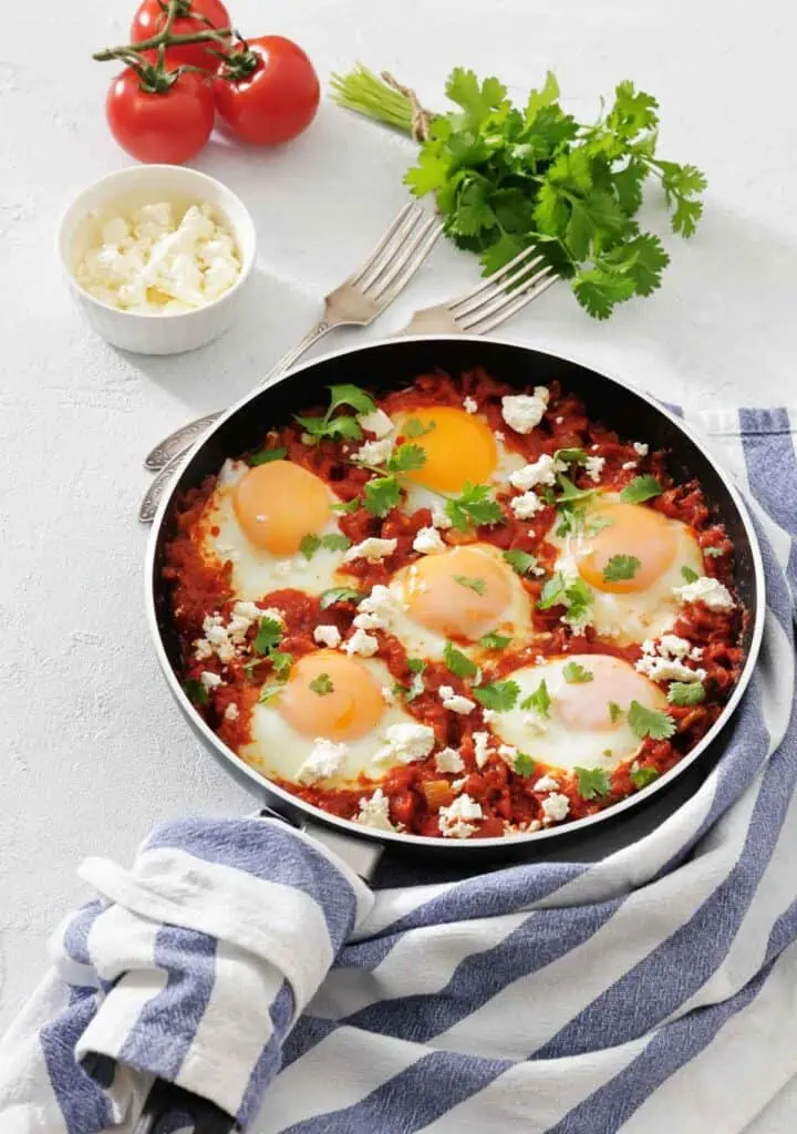 A skillet with eggs poached in a tomato sauce, topped with herbs and cheese. Fresh tomatoes, cilantro, feta, and two forks are nearby on a light surface with a striped towel.