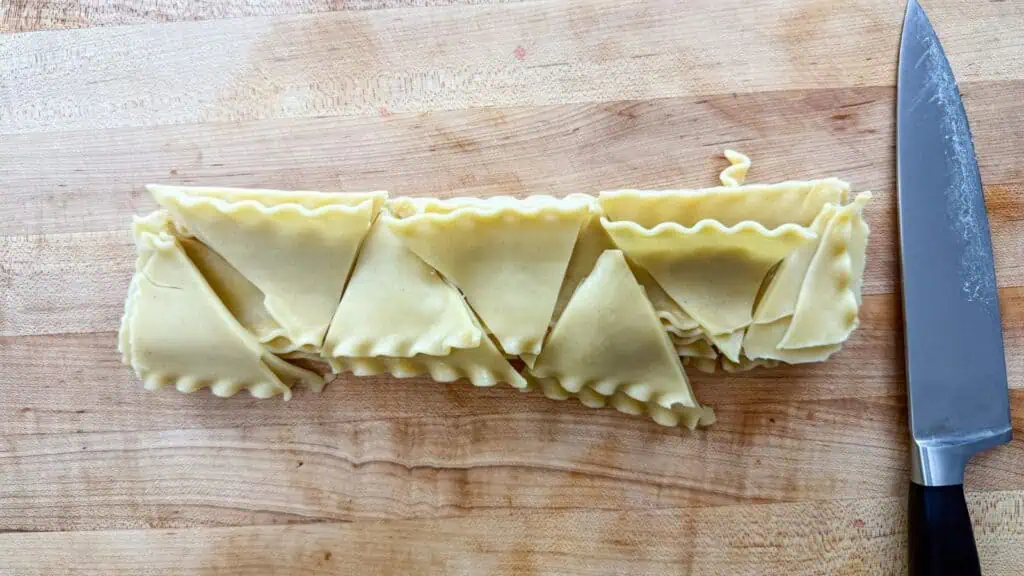 Several uncooked lasagna noodles folded into triangles sit on a wooden surface next to a kitchen knife.