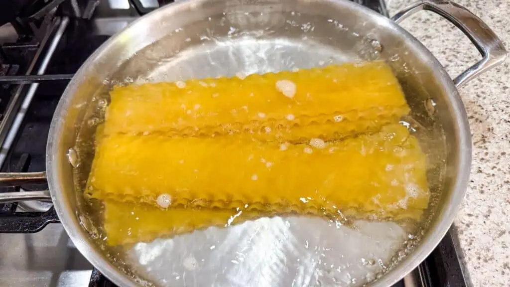 Three lasagna noodles are boiling in a pot of water on a stovetop.