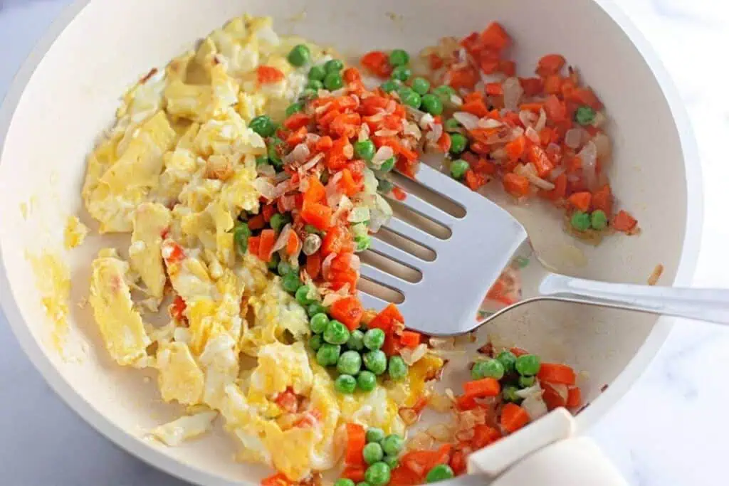 A frying pan with scrambled eggs, sautéed onions, diced carrots, and green peas being mixed with a spatula.