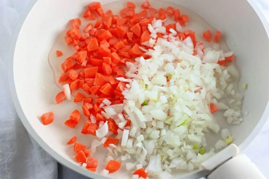 Chopped carrots and onions in a white frying pan with oil, ready to be sautéed.