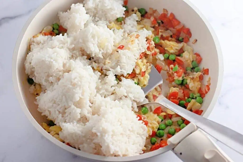 Cooked white rice added to a pan with sautéed chopped carrots, onions, peas, and scrambled egg, all on a white surface with a spatula beside the food.