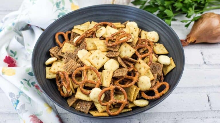 A black bowl filled with snack mix, including pretzels, mini crackers, cereal squares, and round nuts, sits on a white surface next to a floral towel.