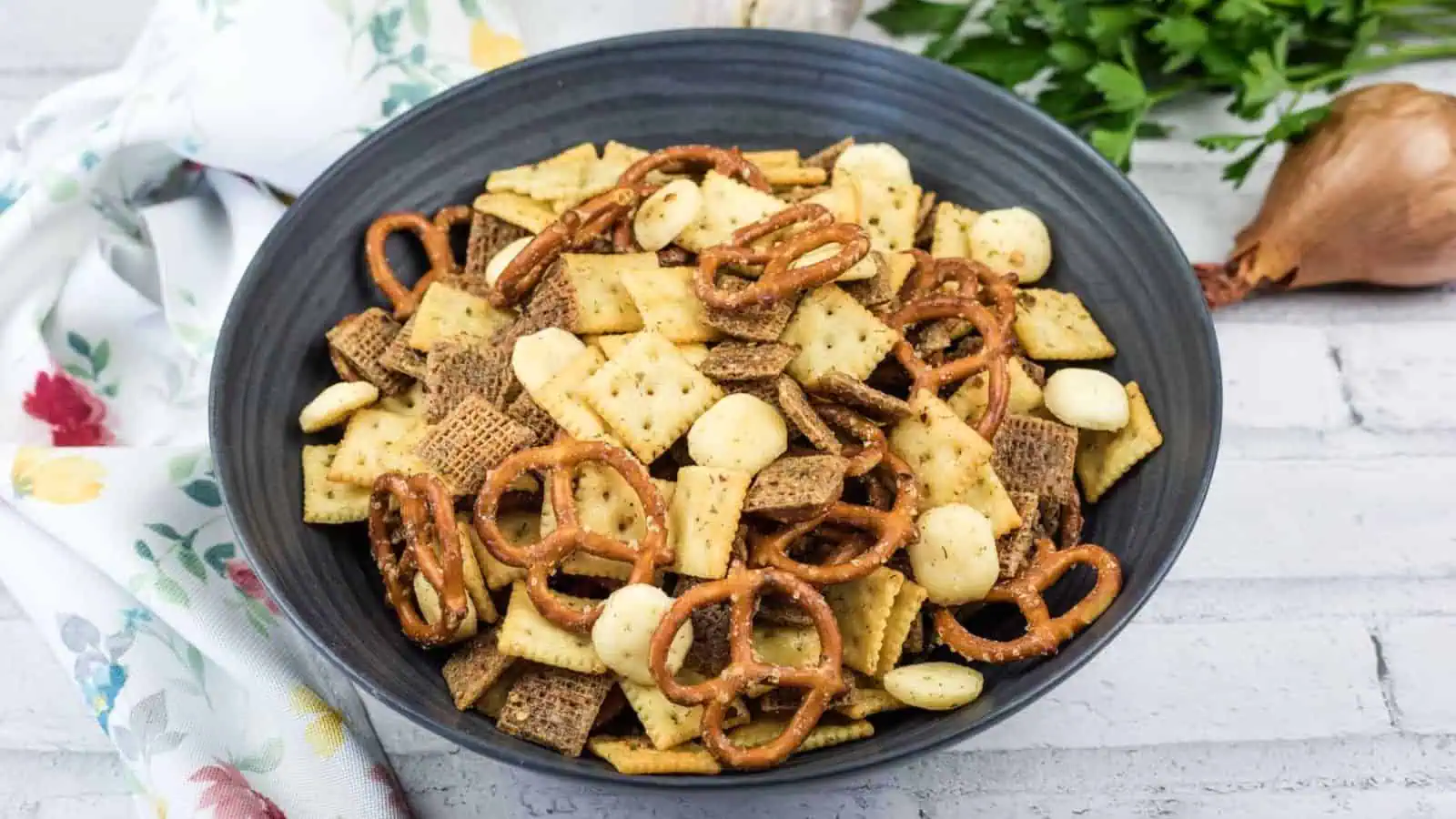 A black bowl filled with snack mix, including pretzels, mini crackers, cereal squares, and round nuts, sits on a white surface next to a floral towel.
