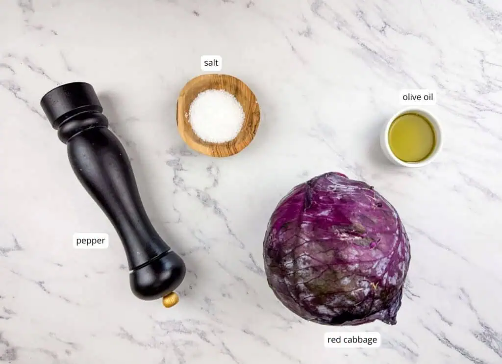 A pepper grinder, a wooden bowl of salt, a cup of olive oil, and a head of red cabbage are arranged on a white marble surface.