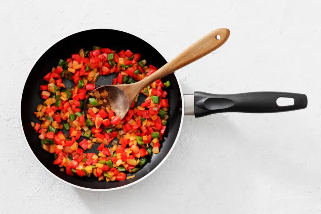 A frying pan with diced red, green, and yellow bell peppers being stirred by a wooden spoon on a white surface.