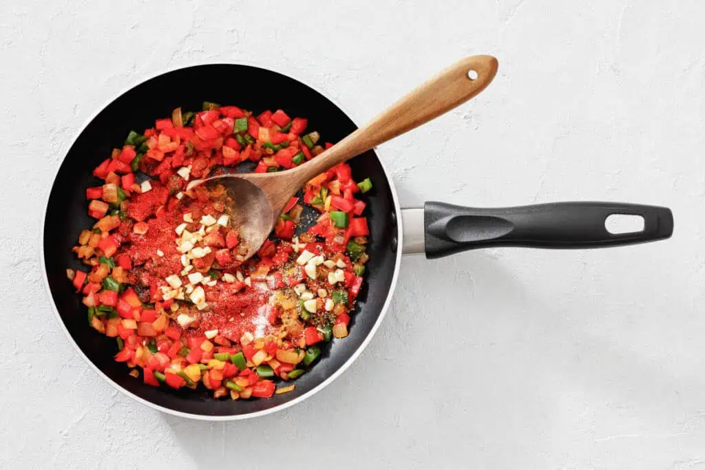 A frying pan with chopped red and green bell peppers, garlic, and spices being stirred with a wooden spoon on a white surface.