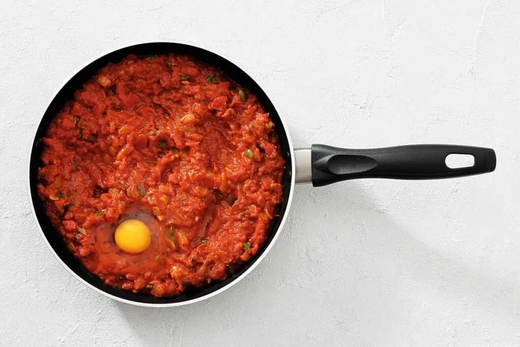 A frying pan with tomato sauce and an uncooked egg in the center, placed on a white surface.